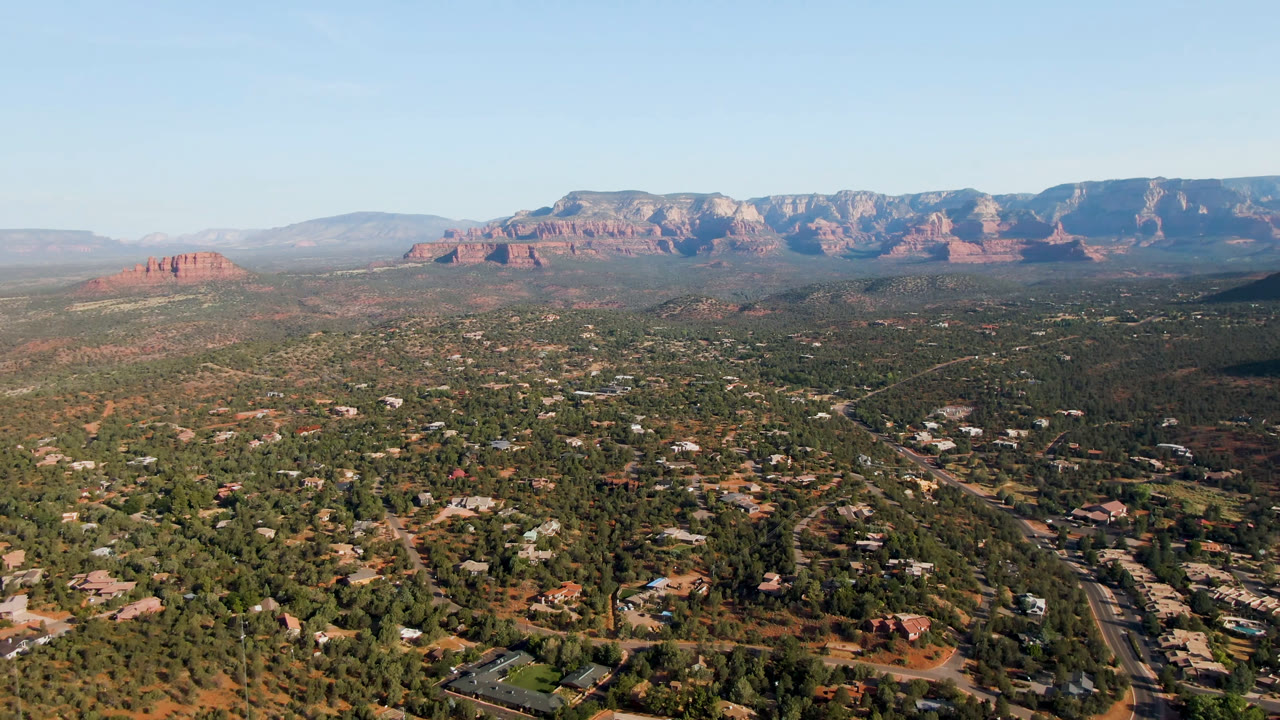 Second-frame aerial over Sedona, Arizona, with more of the road corridor visible in the lower right, scattered homes among trees, red rock ridges, clear sky, and no visible people.