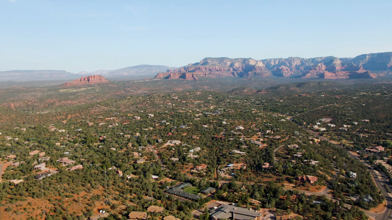 Opening frame of Sedona, Arizona from above, with scattered buildings and trees in the foreground, red rock formations across the horizon, and no visible people.