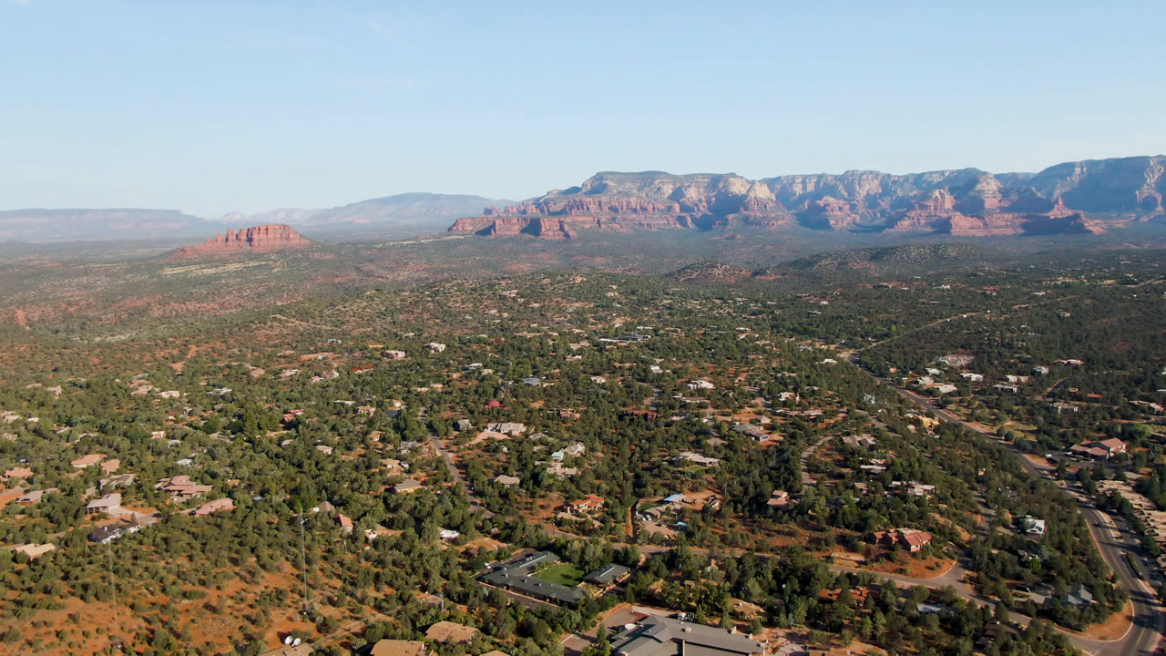 Sedona, Arizona appears in a broad aerial composition, with the road corridor more central, low buildings among trees, distant red cliffs, and no visible people.