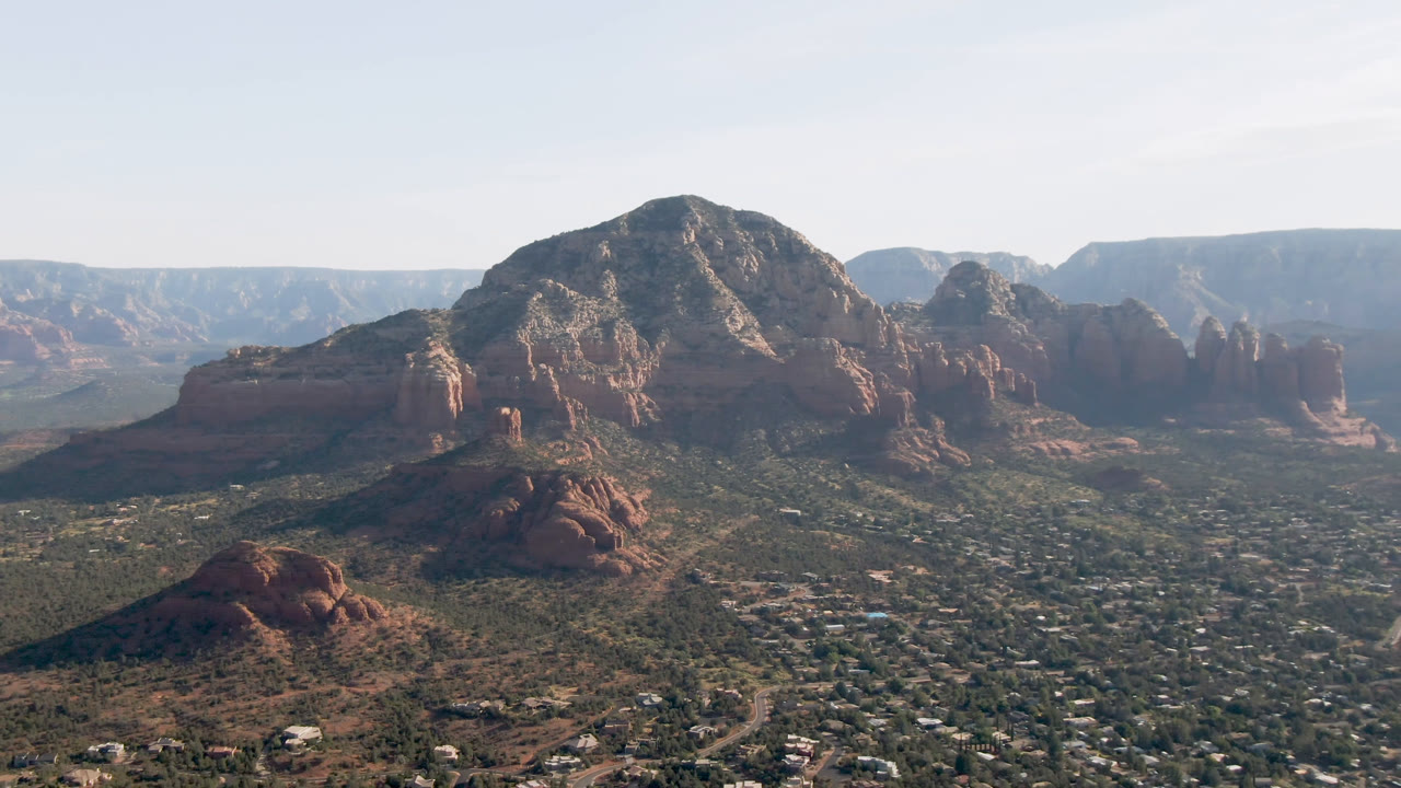 Aerial view across Sedona, Arizona, showing homes and roads in the foreground, red rock formations in the midground, and no visible people.