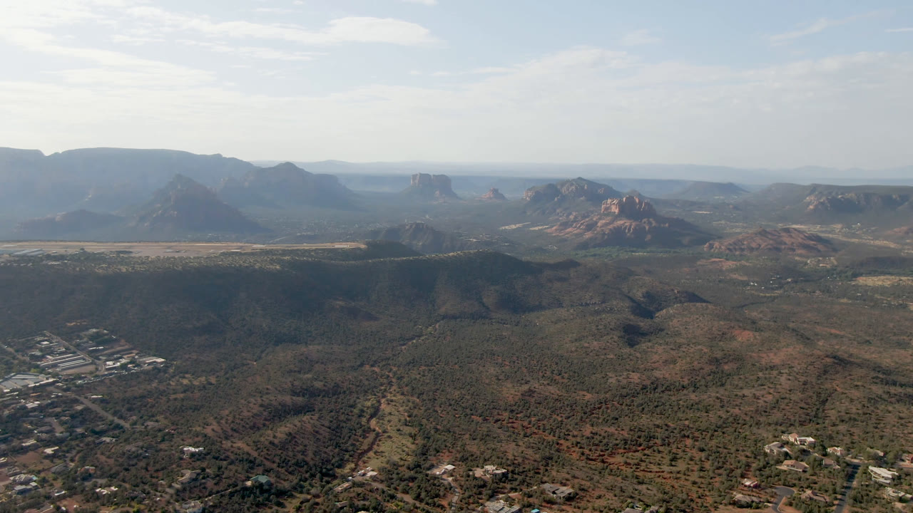 Wide aerial frame over Sedona, Arizona desert valley with red rock formations, distant mesas, pale sky, scattered buildings near the lower edges, and no visible people.