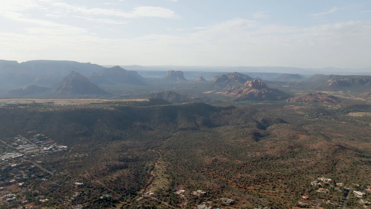 High aerial view of Sedona scrubland and dry washes leading toward red rock buttes, blue-gray distant ridges, light haze, and no visible people.