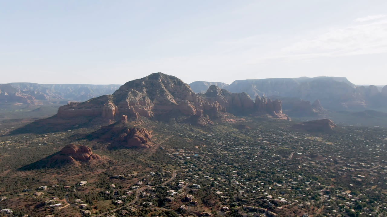 High aerial over Sedona valley with red rock formations, distant mesas, and open sky.