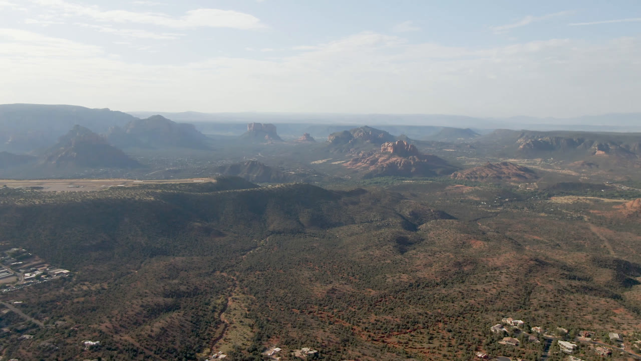Wide aerial view of desert valley, red rock formations, and distant mesas under pale sky.