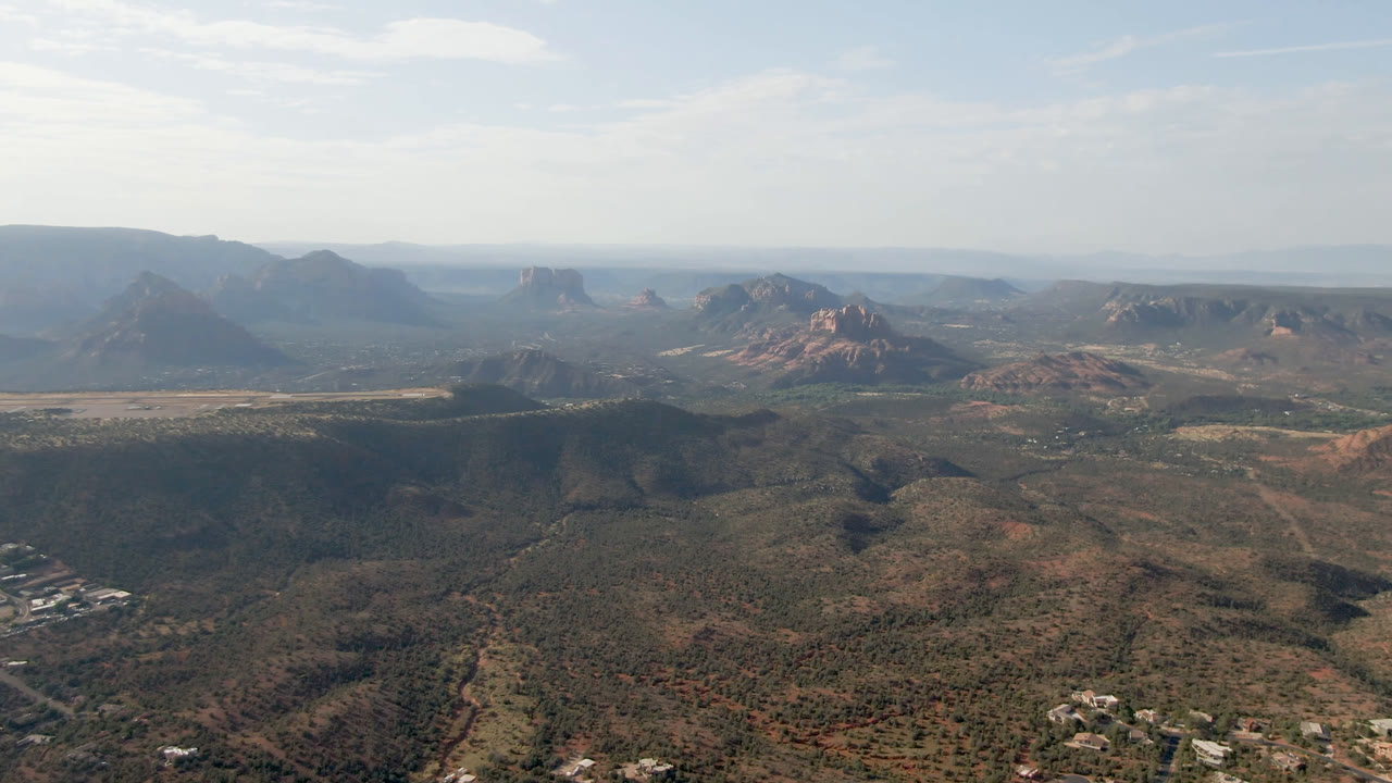 Second frame over Sedona, Arizona showing a wide valley floor below red rock buttes and layered mesas, with small structures near the lower edge and no visible people.