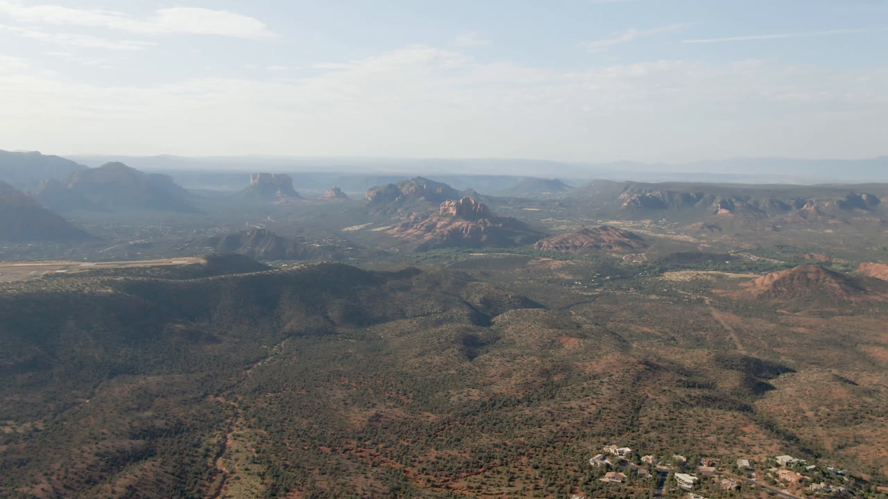Sedona, Arizona aerial frame with a broad desert basin, layered red rock buttes across the middle distance, hazy mesas on the horizon, and no visible people.