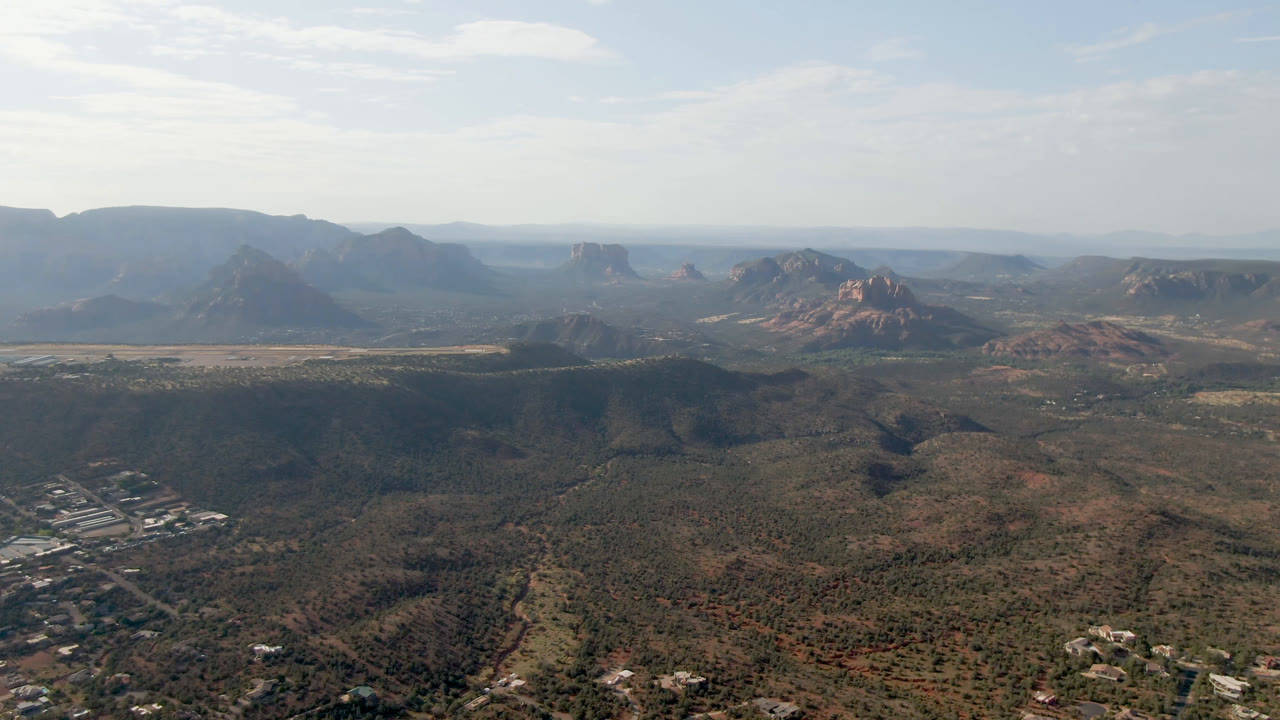 Opening aerial frame over Sedona, Arizona, with scrub-covered valley ground, scattered town buildings at lower left, red rock formations in the distance, and no visible people.