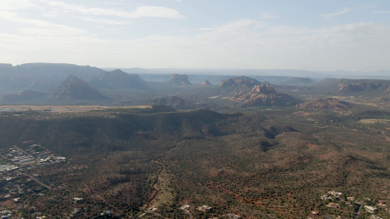 One second into the Sedona aerial, the frame holds a wide valley composition with town structures at lower left, red rock buttes across the horizon, and no visible people.