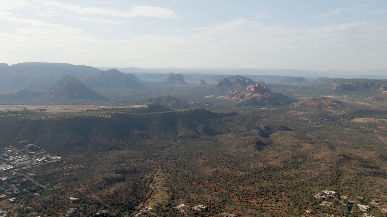 Aerial view across scrubland toward red rock formations and broad mesa silhouettes.