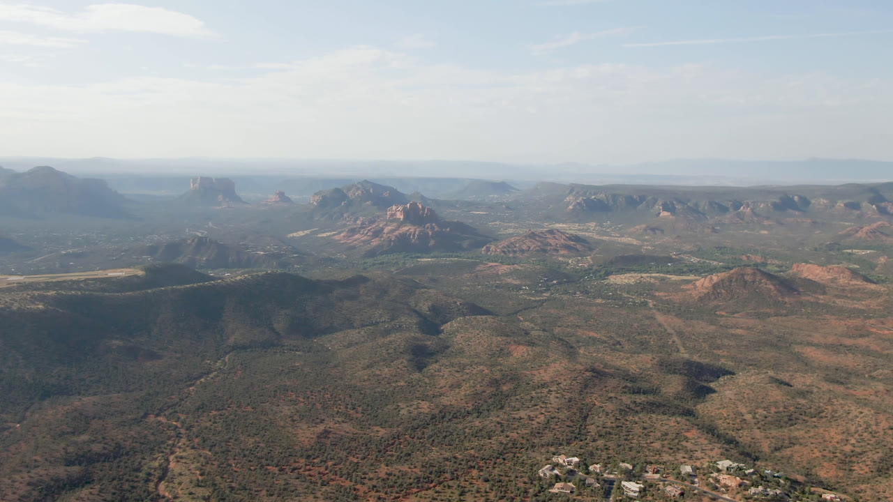Wide aerial view of desert hills, red rock formations, and a pale blue sky.