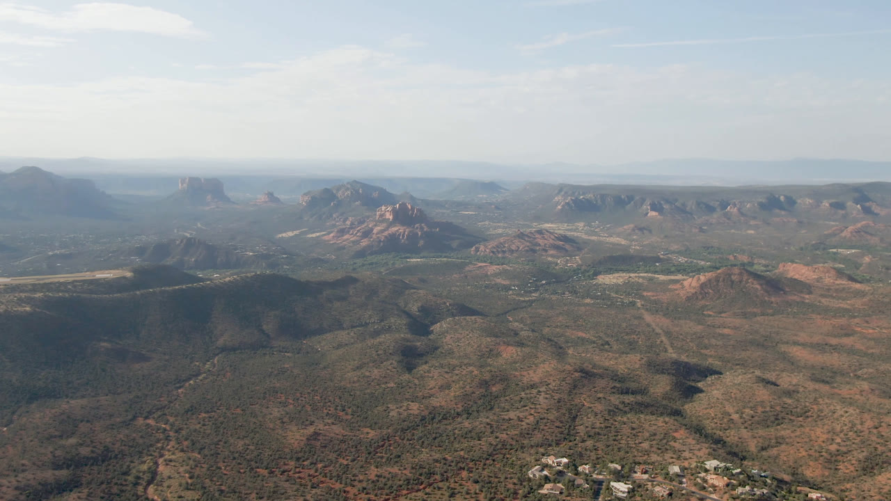 Second aerial frame over Sedona, Arizona, looking across darker foreground ridges toward red rock peaks, open valley floor, hazy distant mesas, and no visible people.