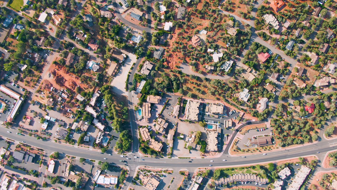 Overhead frame of Sedona, Arizona showing a curving main road, side streets, scattered buildings, red dirt, trees, and small vehicles with no visible people.
