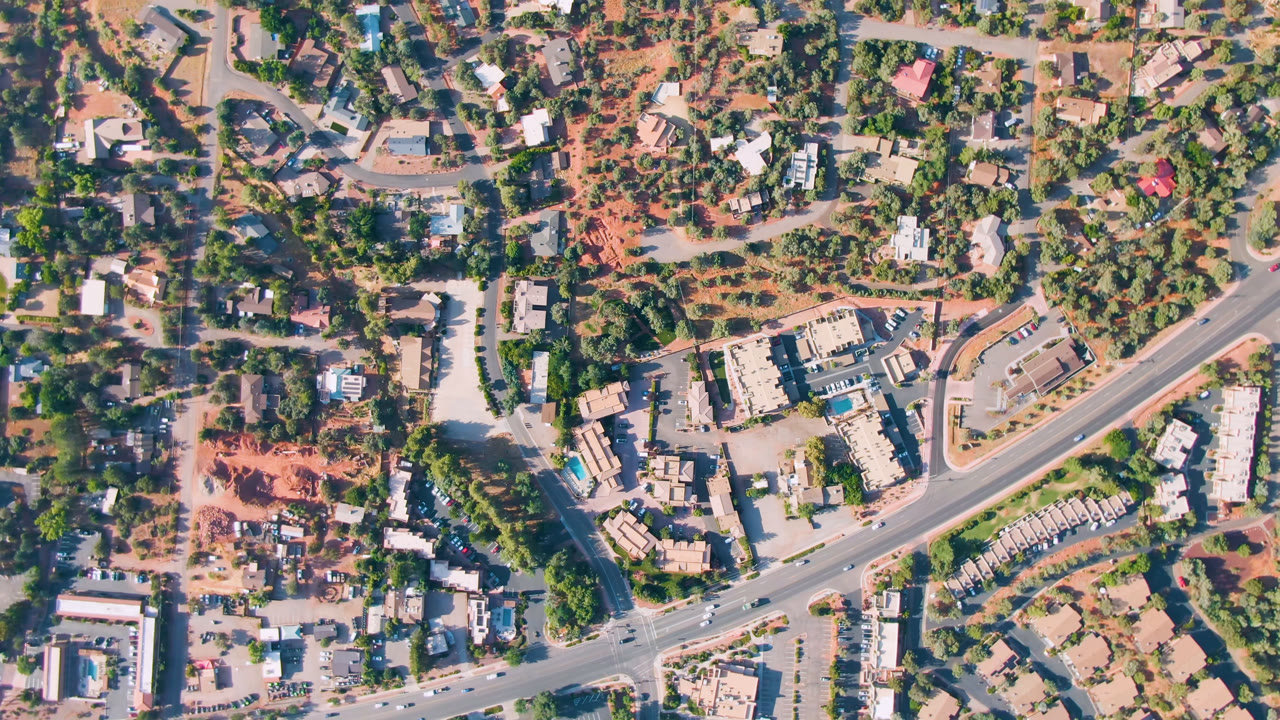 Birdseye Sedona, Arizona view with the main road, a curved side street, clusters of homes, green vegetation, open dirt areas, vehicles, and no visible people.
