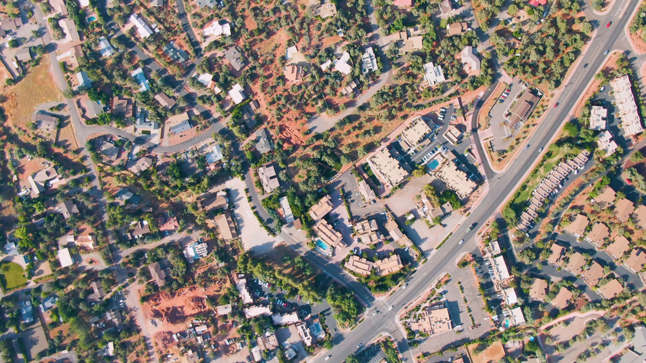 Sedona aerial frame with a central roadway, clustered rooftops, parking lots, desert-colored ground, tree-lined blocks, and no visible people.
