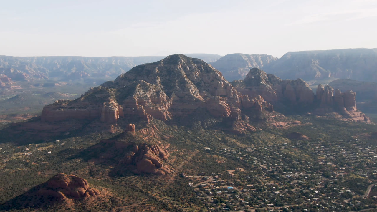 Aerial view in Sedona, Arizona, with a central red rock mass, scattered rooftops on the valley floor, layered cliffs in the distance, and no visible people.