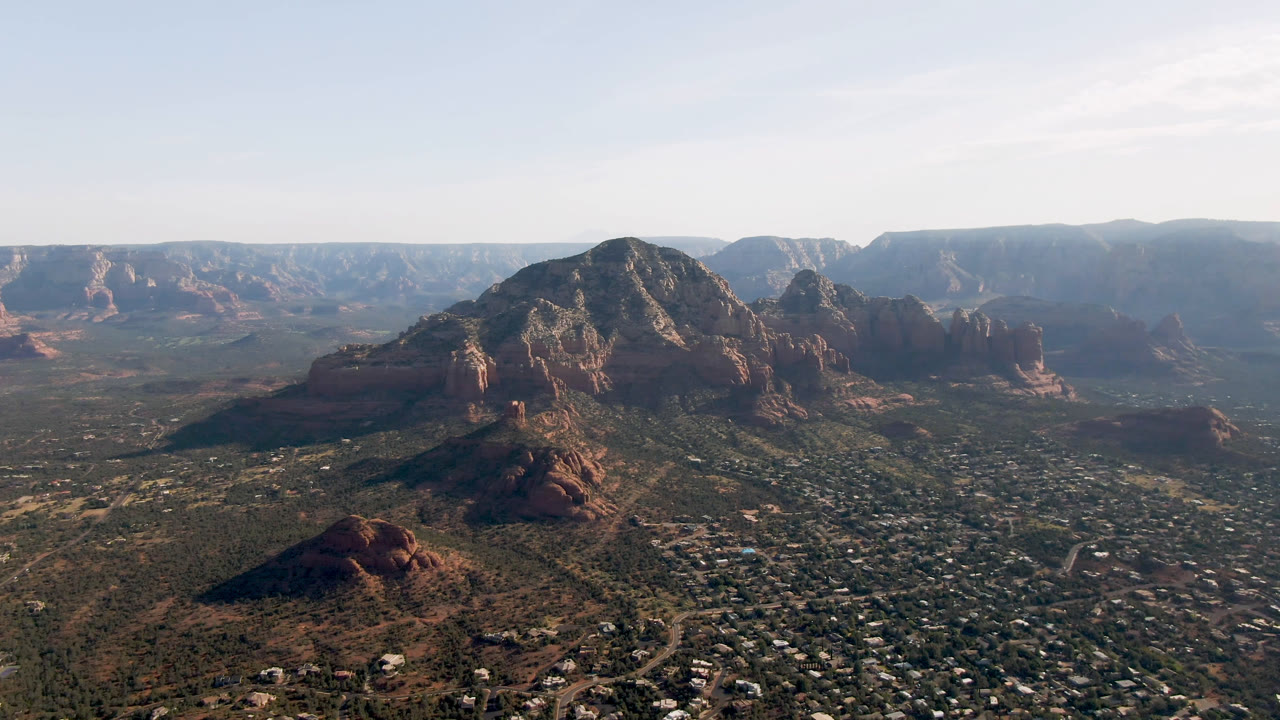 Second frame of the Sedona aerial, centered on a large shadowed red rock massif with scattered buildings and roads across the valley floor, and no visible people.