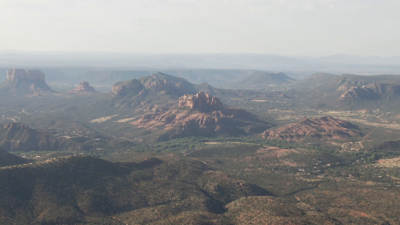 Opening frame of Sedona, Arizona from high above, showing red rock formations beyond scrub hills, a broad valley floor, distant plateau lines, and no visible people.