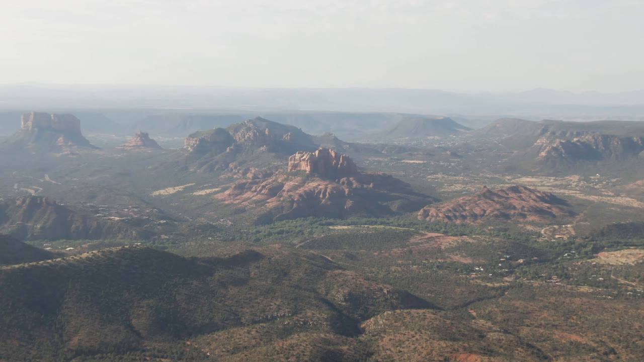 High aerial frame over Sedona, Arizona with a central red rock bluff, darker foreground ridges, muted green desert vegetation, hazy distance, and no visible people.