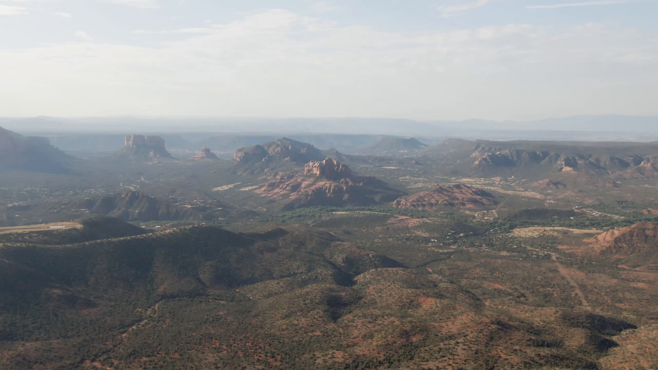 Wide aerial view of red rock hills and a desert valley under thin clouds.