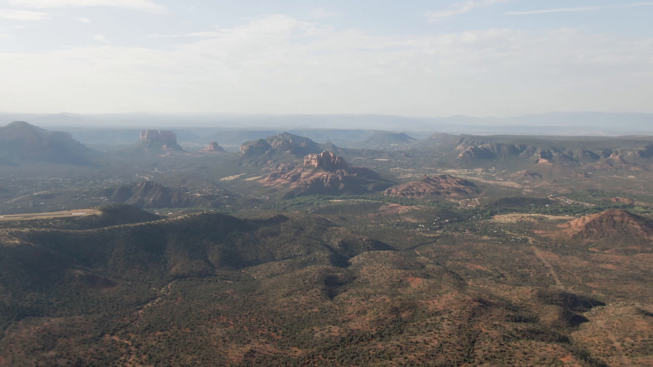 Second frame of the Sedona aerial holds on red and brown desert ridges, scattered midground formations, a long valley floor, hazy mesas, and no visible people.