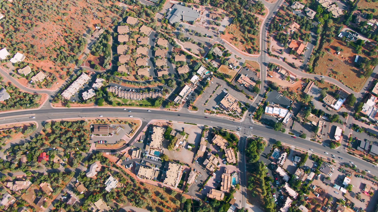 Overhead aerial frame of Sedona, Arizona showing a paved road, side streets, scattered buildings, trees, reddish open ground, and no visible people.