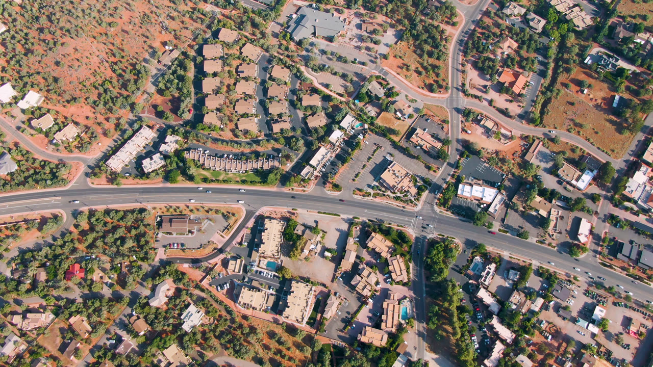 High aerial frame over Sedona, Arizona with a main road running across the lower area, a curved side street, clustered buildings, trees, and no visible people.