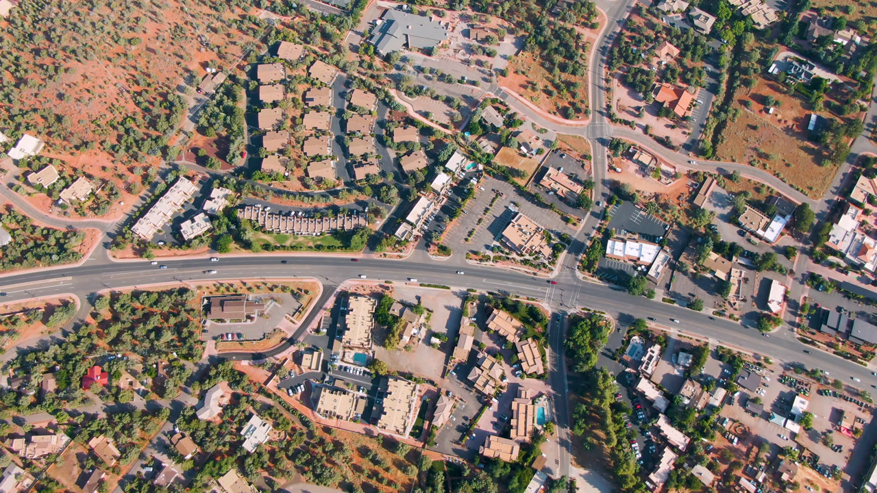 High aerial frame over Sedona, Arizona, showing a curving main road, side streets, low buildings, trees, red open ground, vehicles, and no visible people.