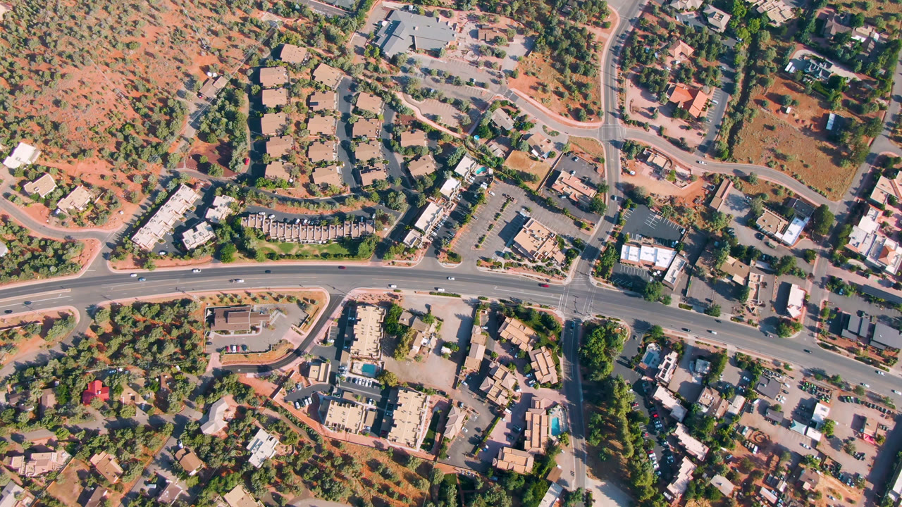 Overhead Sedona, Arizona frame with the main road running across the lower area, clustered buildings and parking lots near the center, red terrain above, vehicles, and no visible people.