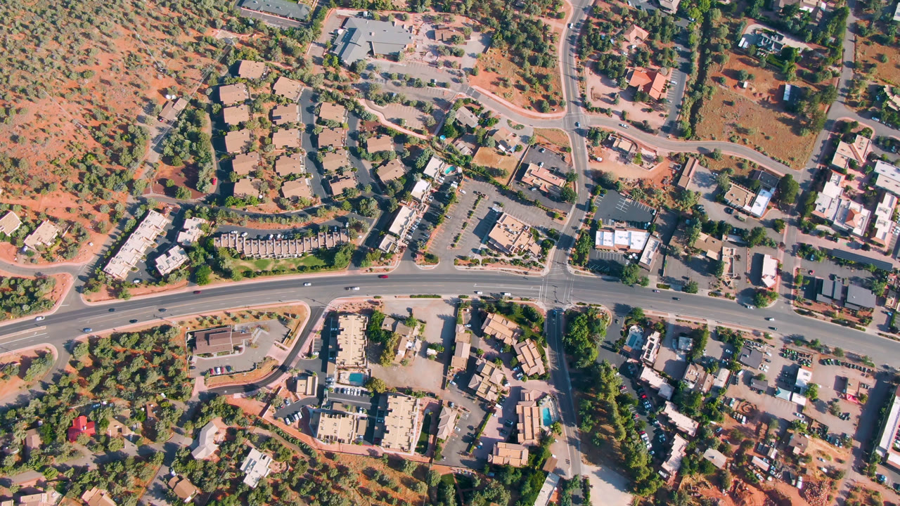 Second overhead frame in Sedona, Arizona with the main road running across the lower half, side streets, clustered buildings, tree cover, red ground, and no visible people.