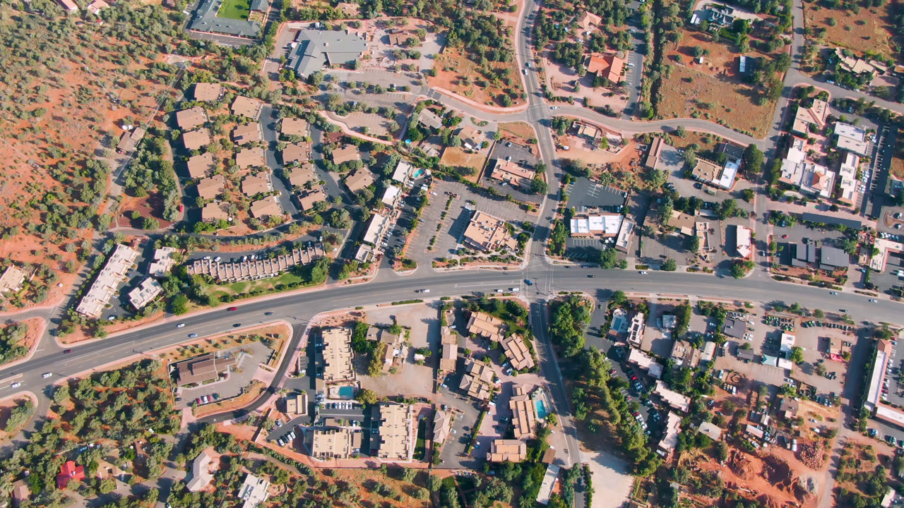 Opening overhead frame of Sedona, Arizona with a curved main road, side streets, low buildings, parking lots, trees, red earth, and no visible people.