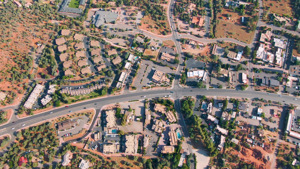 Second overhead frame of Sedona, Arizona centered on a multi-road junction, roadside buildings, parked and moving cars, trees, red ground, and no visible people.