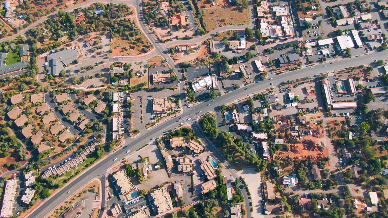 Second overhead frame of Sedona, Arizona showing the main road running across the upper frame, smaller streets, parking areas, homes or low buildings, trees, and no visible people.