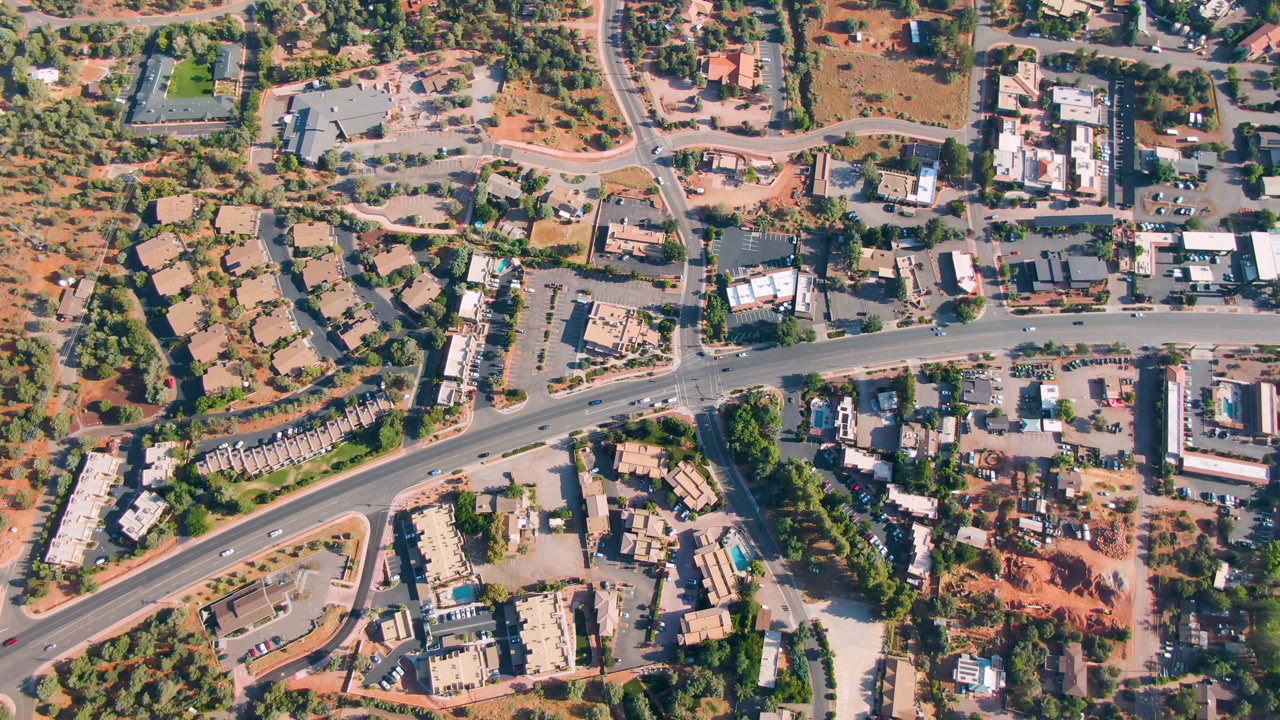 Overhead Sedona frame with the main road crossing the center, clusters of rooftops on both sides, scattered vehicles, trees, and reddish desert terrain, with no visible people.