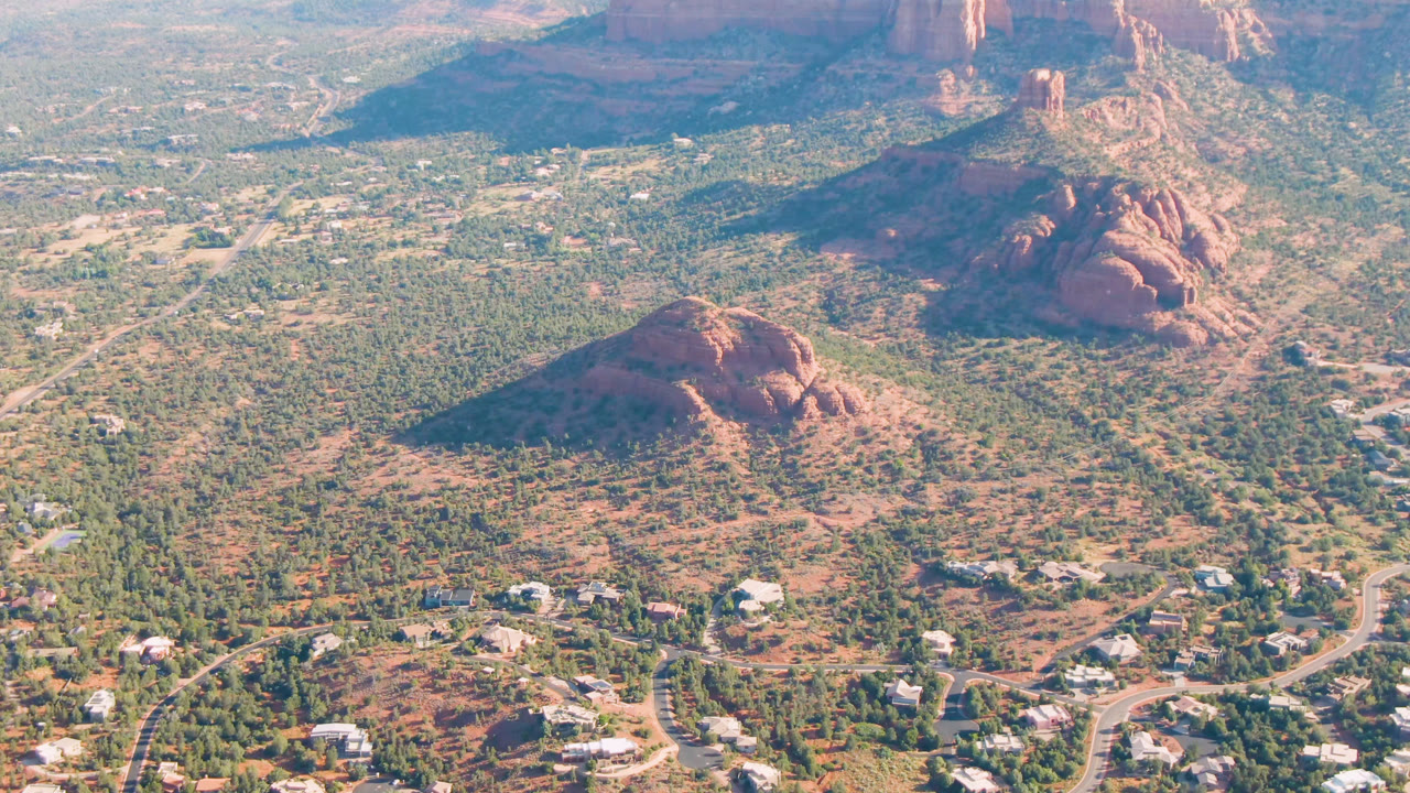Opening aerial frame over Sedona, Arizona, showing scattered buildings, winding roads, green desert scrub, red rock hills, distant cliffs, and no visible people.