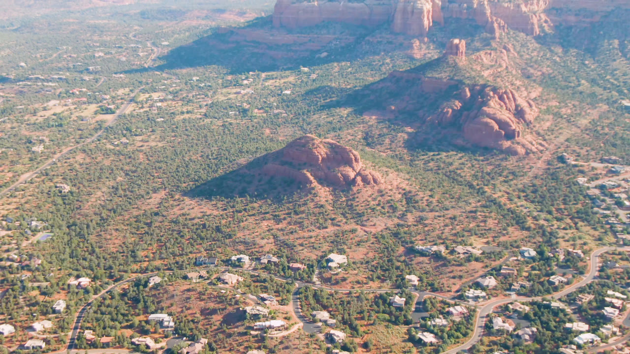 Early aerial frame in Sedona, Arizona, with a rounded red hill near center, residential roads below, tall cliffs in the distance, and no visible people.