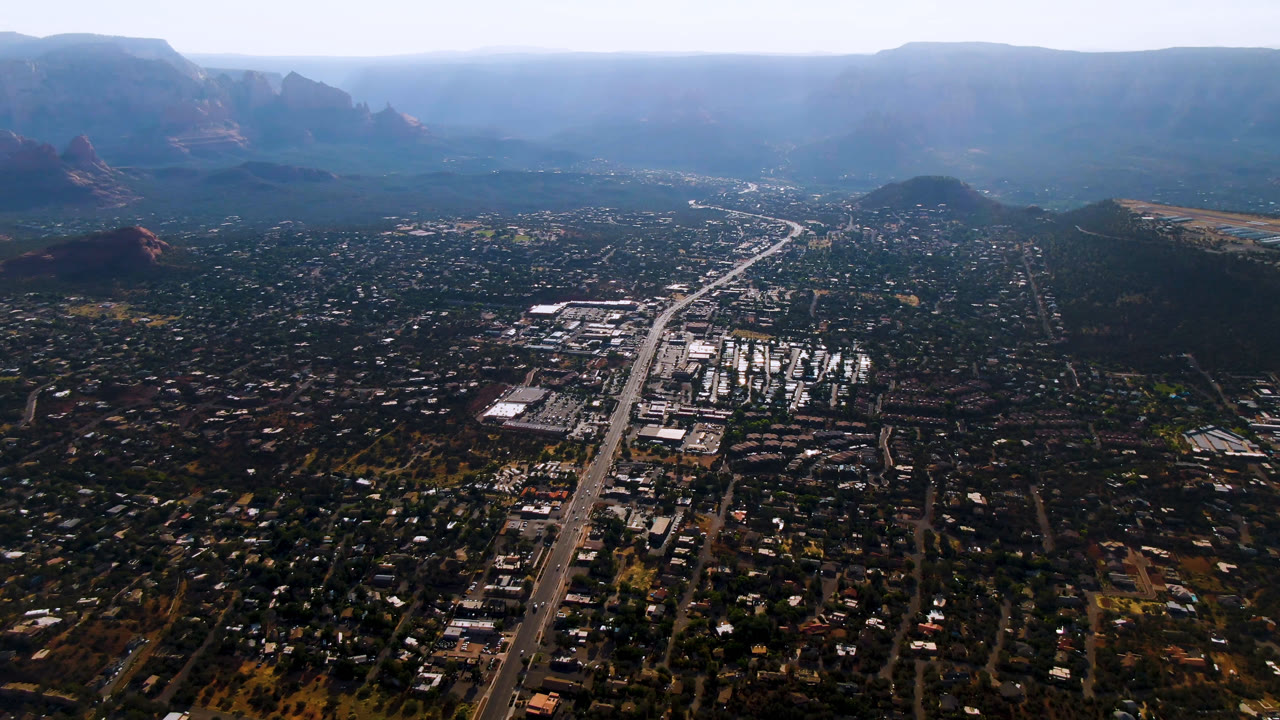 Opening frame of Sedona, Arizona from high above, with a straight highway through the town, red rock hills at the edges, a hazy mountain horizon, and no visible people.