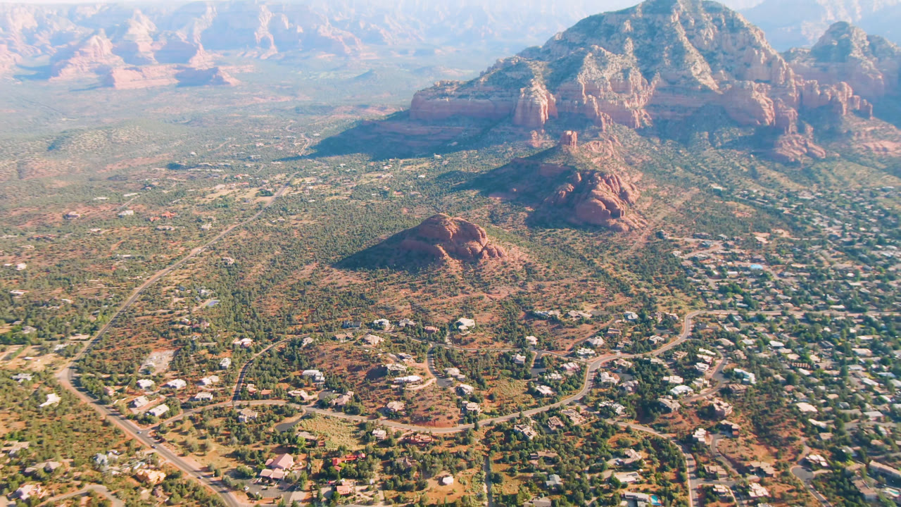 High aerial view over Sedona, Arizona, with a central red rock butte, neighborhoods and roads below, mountain formations across the background, and no visible people.