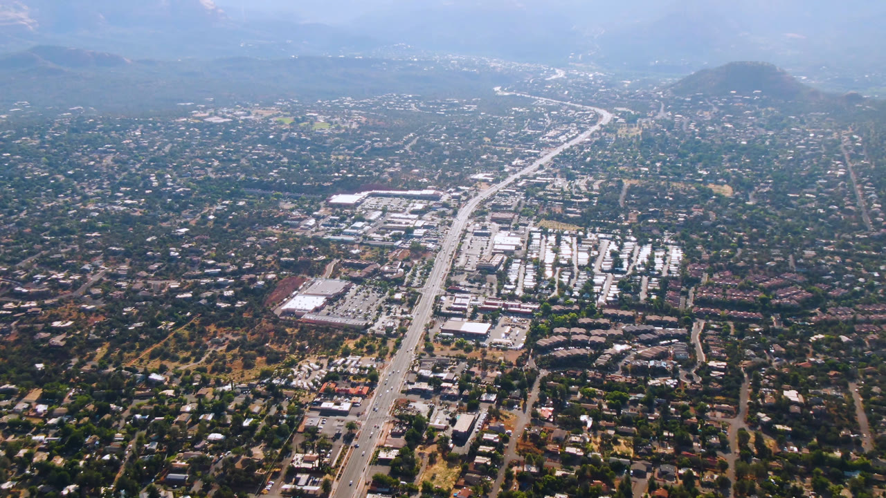 Second aerial frame of Sedona, Arizona, showing more of the central highway corridor, clustered white-roof buildings, surrounding neighborhoods, hazy hills, and no visible people.