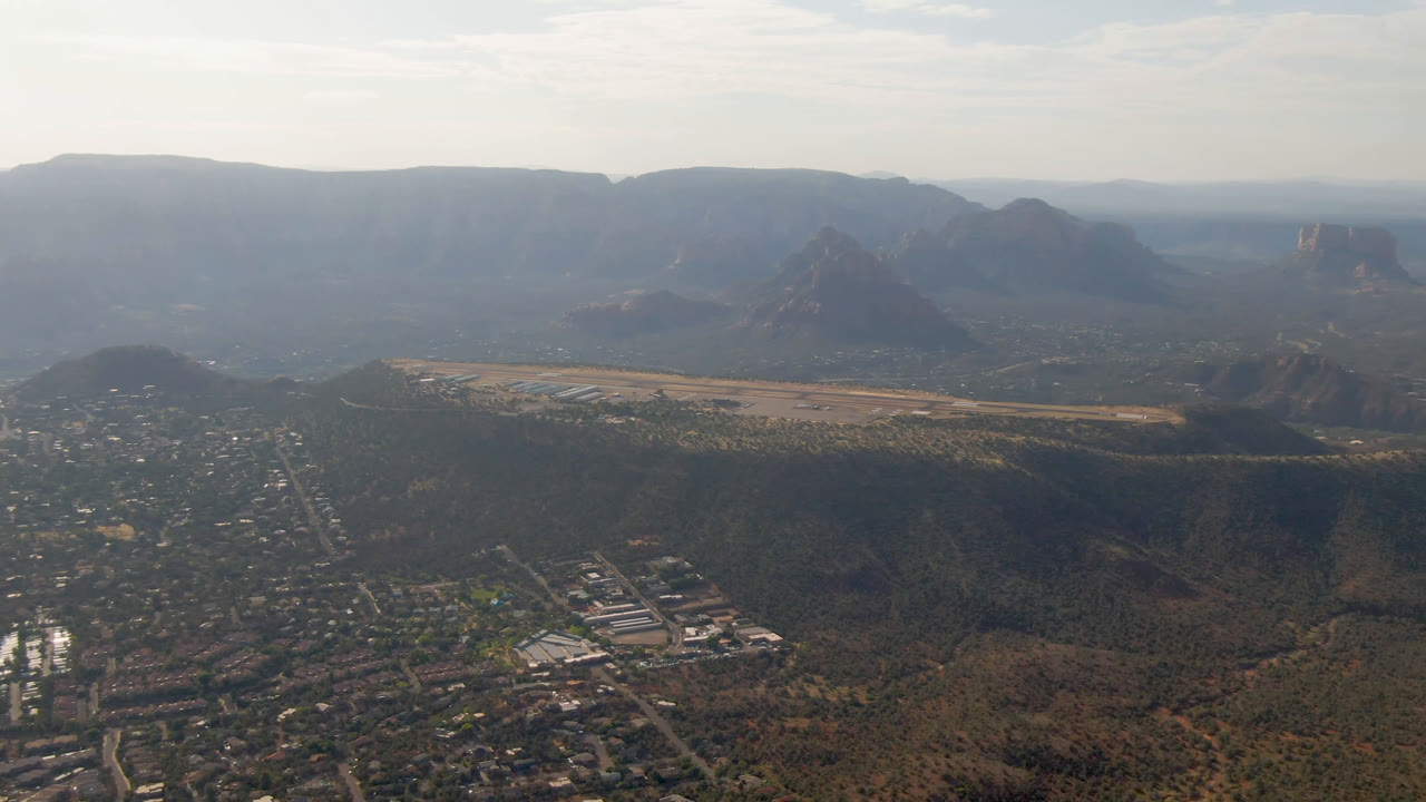 Sedona, Arizona appears from a high aerial angle with clustered buildings at lower left, a dark ridge across the midground, distant rock formations, and no visible people.