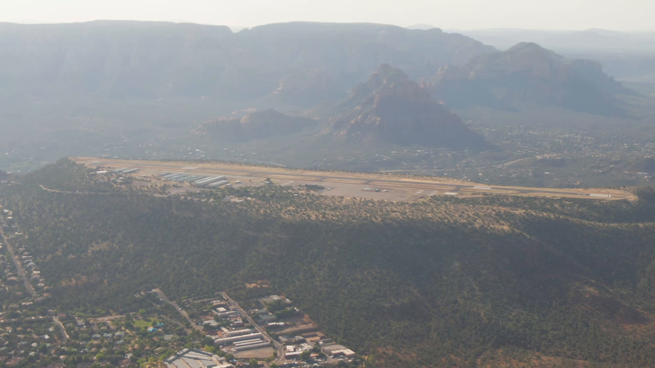 One second in, the Sedona aerial holds on a central road corridor, scattered rooftops, a pale open strip, dark green hills, distant red rock peaks, and no visible people.
