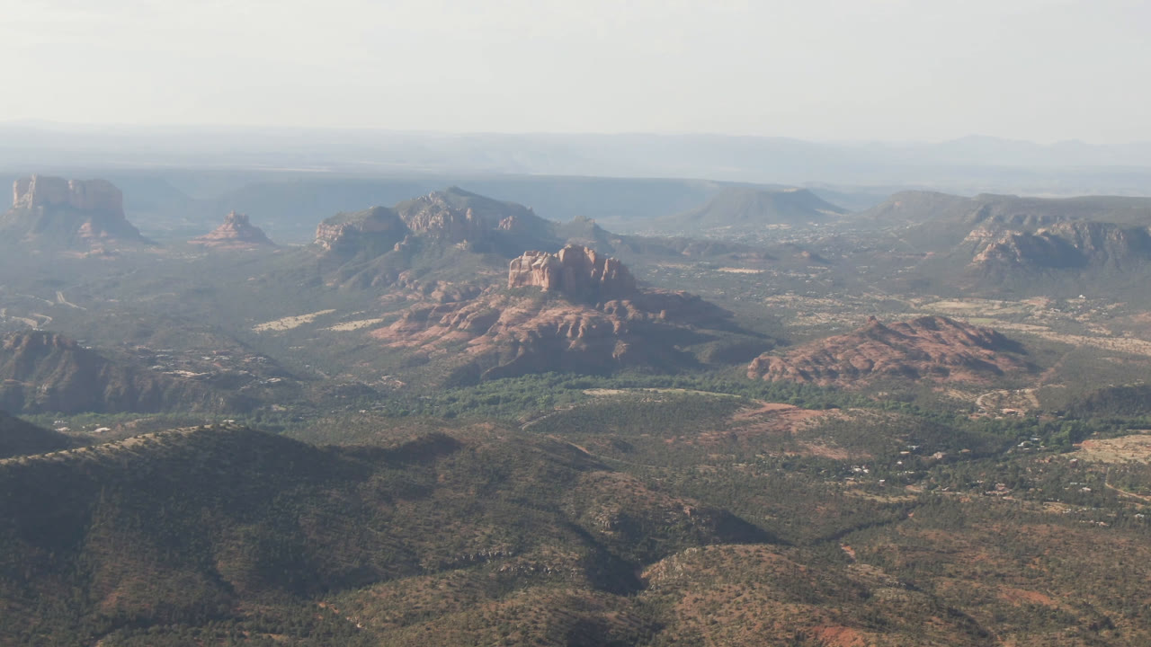Opening aerial frame over Sedona, Arizona shows scrub-covered foreground hills, red rock formations in the distance, layered horizon haze, and no visible people.