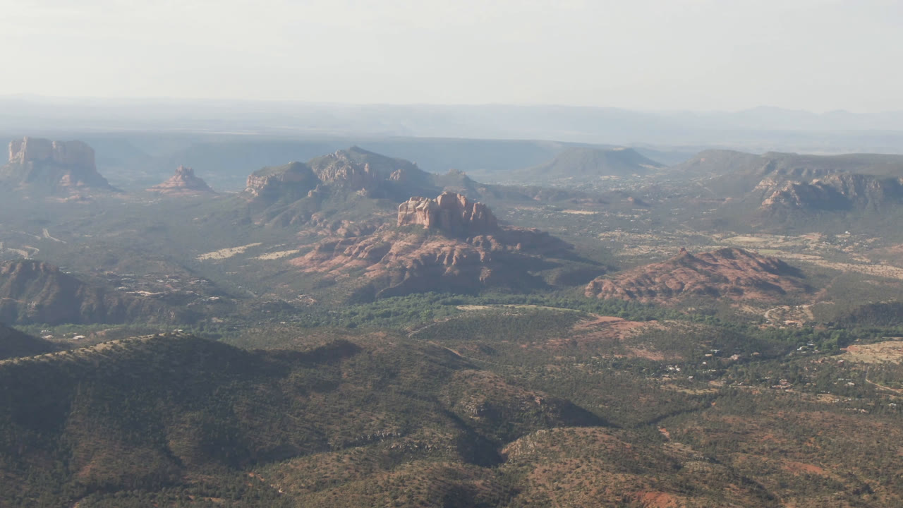 At one second, the Sedona aerial view holds on low desert hills in the foreground, red ridges and buttes beyond, a broad valley floor, and no visible people.