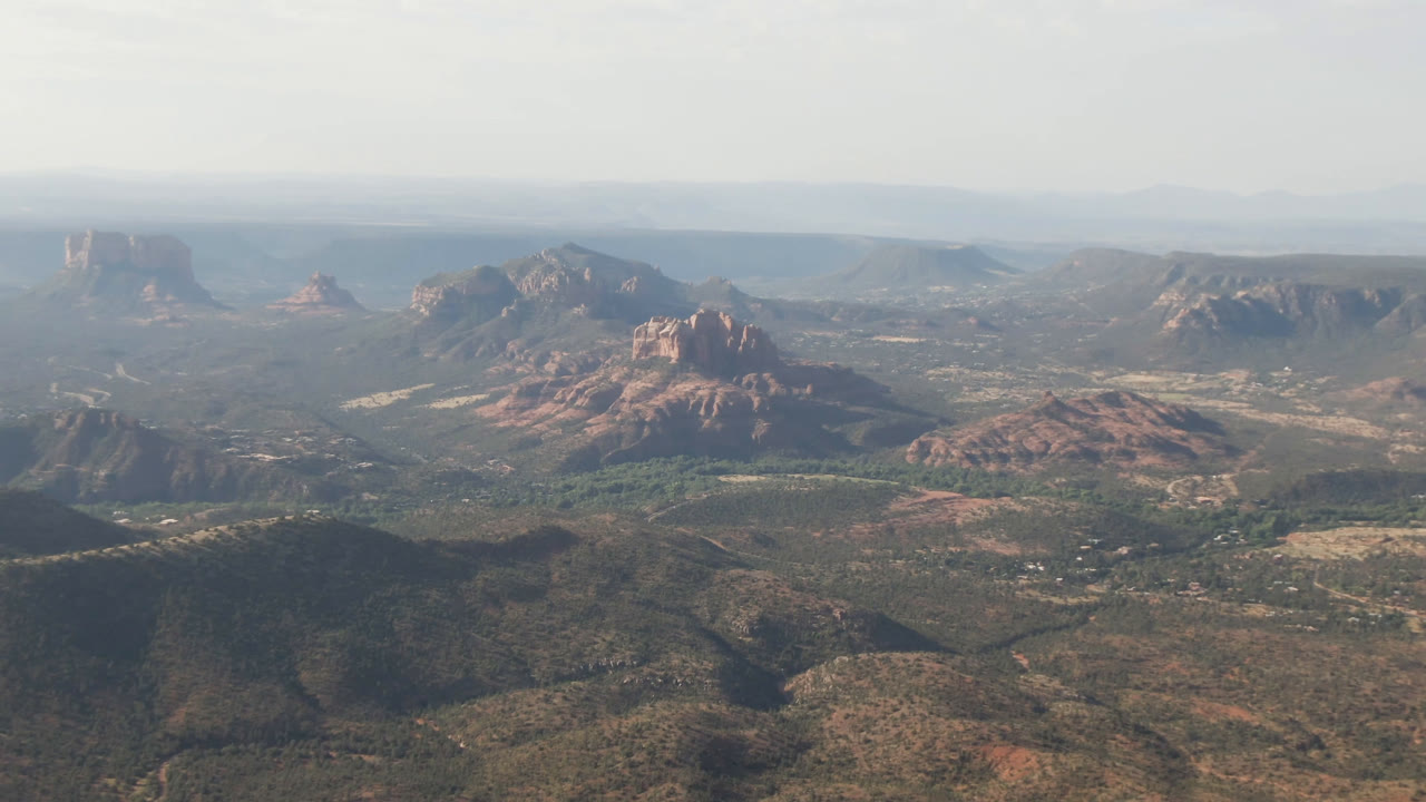 High aerial view across scrub hills toward red rock mountains and mesas.
