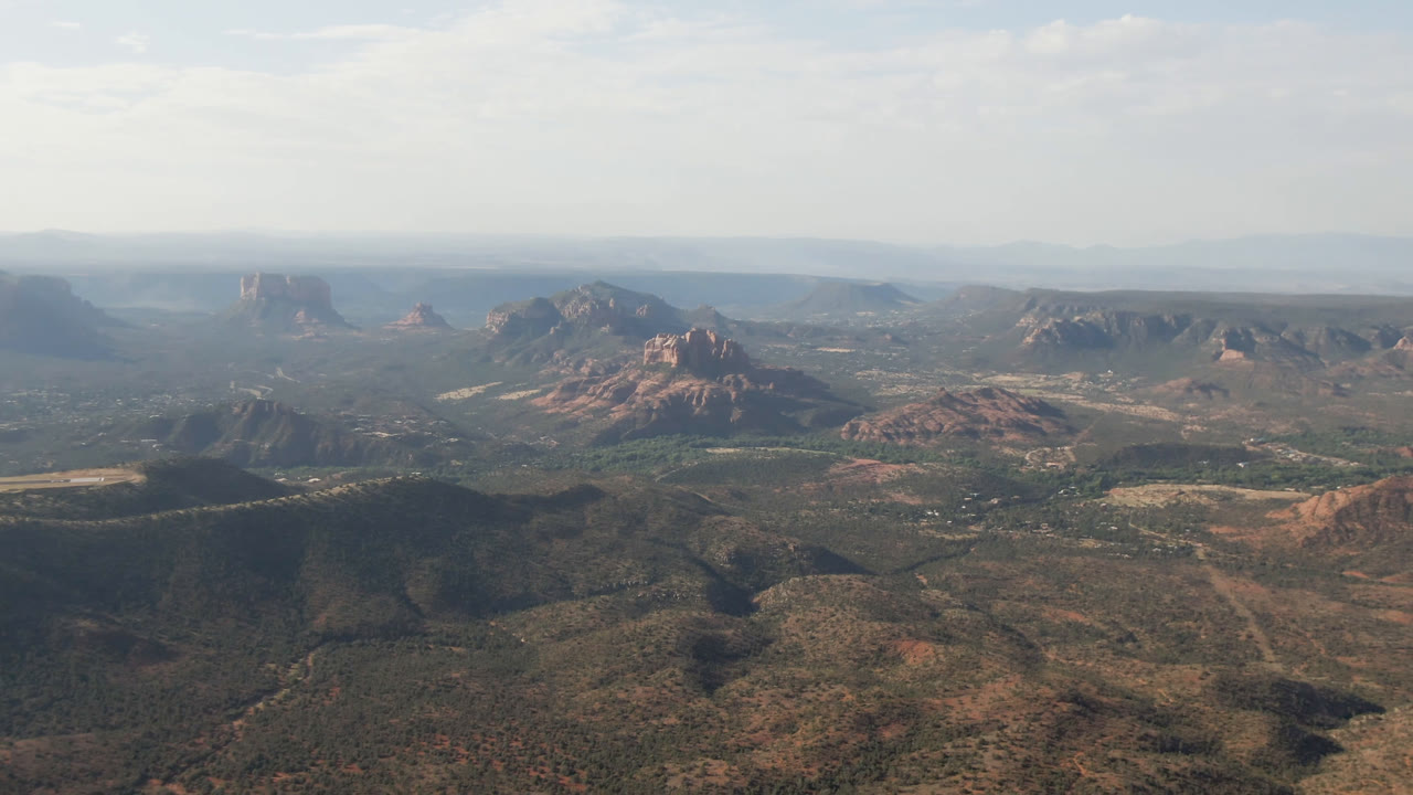 Opening aerial frame over Sedona, Arizona, showing scrub-covered valley floor, red rock ridges, distant mesas, thin clouds, and no visible people.