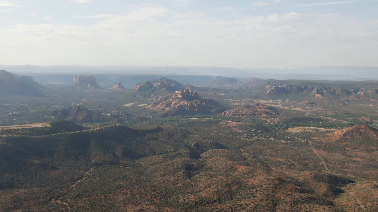 Aerial frame over Sedona, Arizona, with dark scrubby hills in the foreground, an open valley midground, red rock buttes near center, hazy mountain distance, and no visible people.