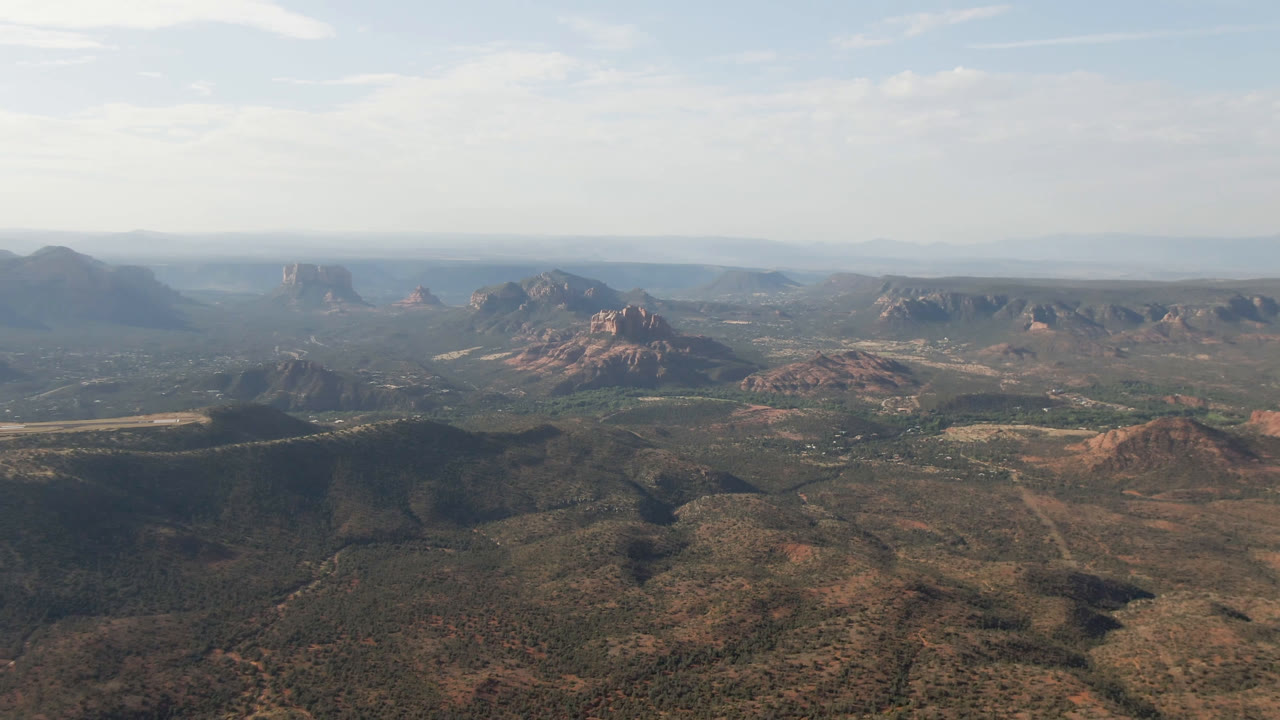 Aerial view across scrub desert toward red rock ridges and distant mesas beneath light cloud cover.