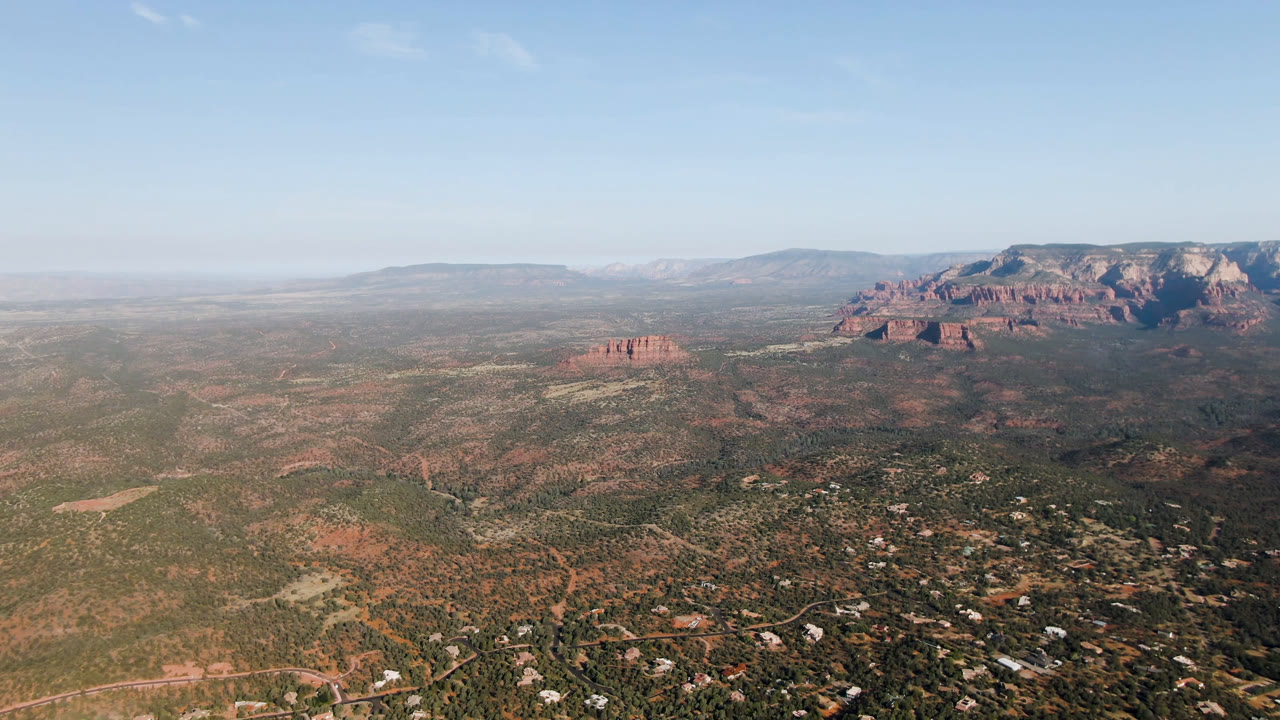 Opening aerial frame over Sedona, Arizona, with desert scrub, scattered valley buildings, red rock formations on the right, distant mesas, pale blue sky, and no visible people.