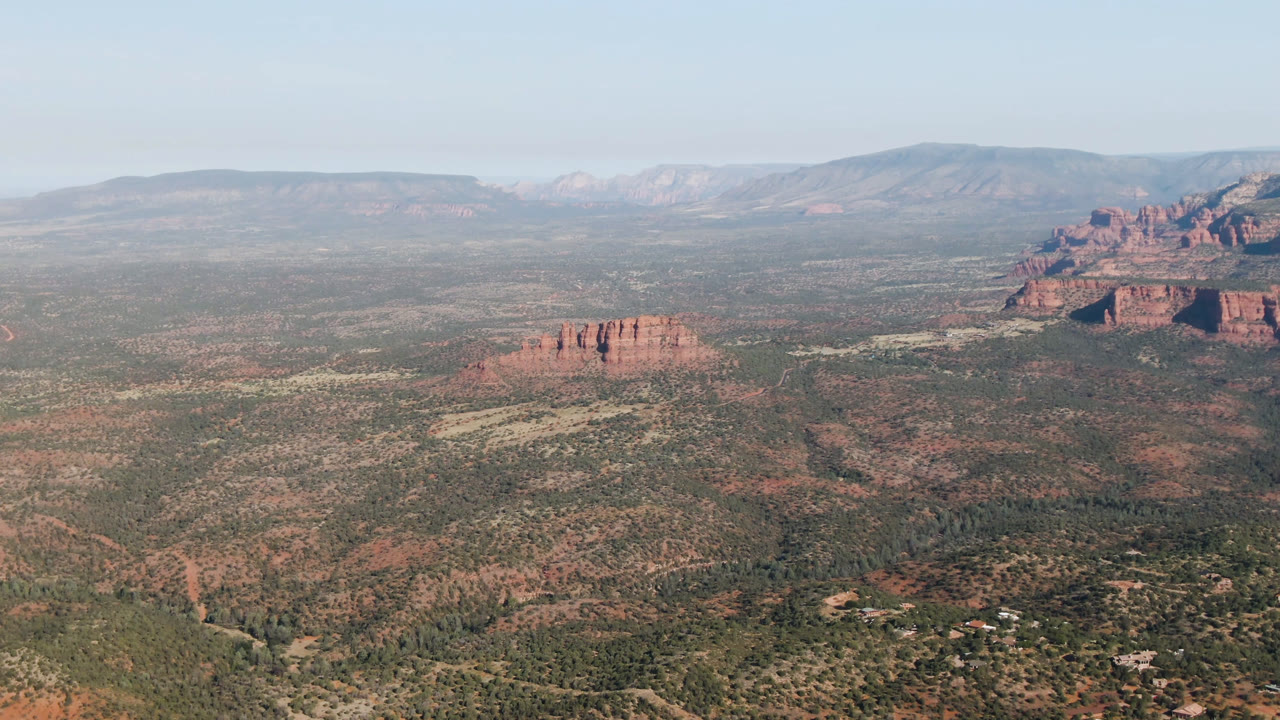 Opening frame of Sedona, Arizona from an elevated wide view, with brushy valley floor, red rock formations, hazy distant ridges, pale blue sky, and no visible people.