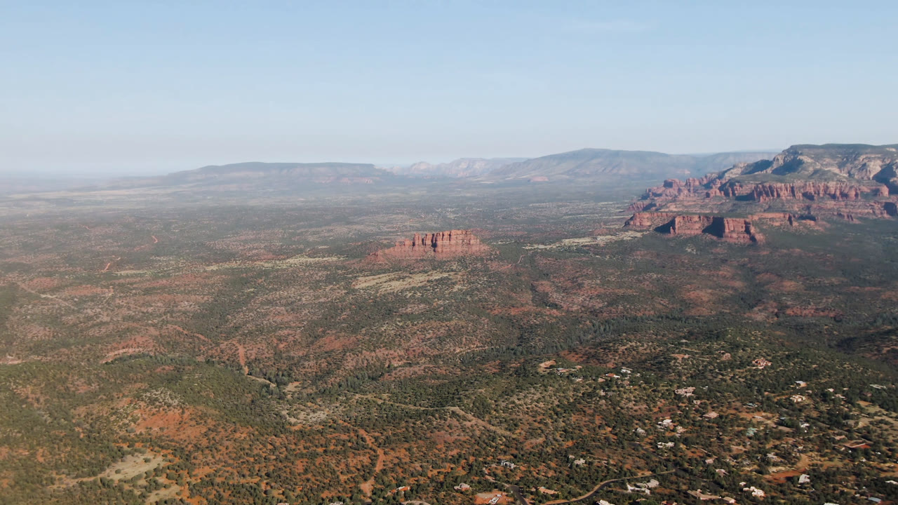 Wide aerial view of desert valley, red rock formations, distant mesas, and blue sky.