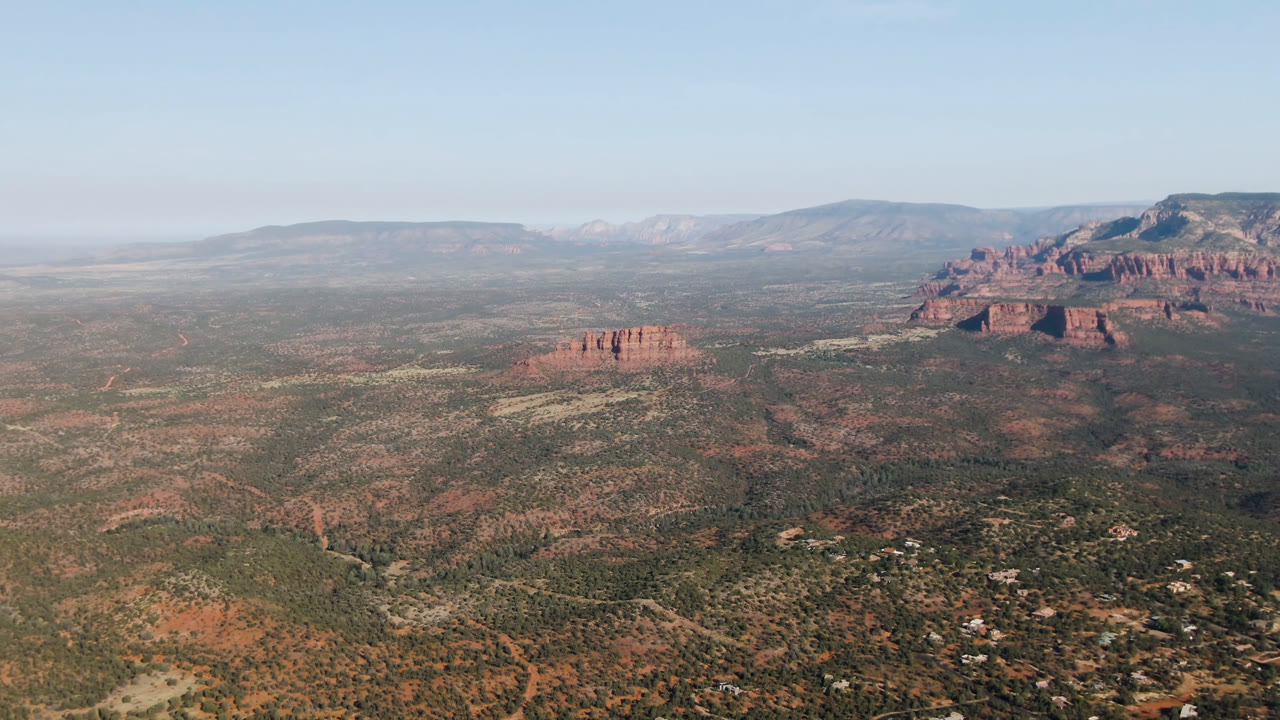 Aerial view in Sedona, Arizona with an open desert valley below, a red rock bluff mass on the right, a smaller butte left of center, distant mesas, pale daylight, and no visible people.